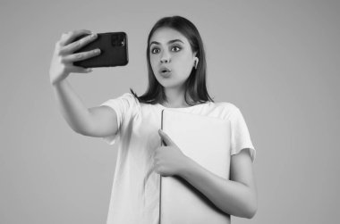 Beyaz tişörtlü, elinde telefon ve dizüstü bilgisayarla gri arka planda izole edilmiş genç bir bayan öğrenci. Öğrenci video görüşmesi yapıyor. Stüdyoda akıllı telefonu olan kız öğrenci. Video çağrısı alan öğrencinin portresi