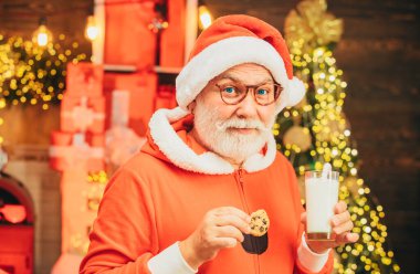 Noel Baba, Noel arifesinde kendisine bırakılan kurabiye ve sütü sever. Neşeli Noel Baba elinde sütle bardak, şömineli kurabiye ve arka planda Noel ağacı tutuyor.