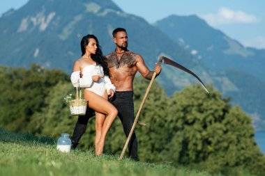 Çiftlikte sıcak yaz işleri. Yeşil çimen tarlasında sarılan seksi kadınla güçlü üstsüz çiftçi. Kaslı çiftçi yaz otlağında karısını kucaklıyor. Yaz çiftçileri