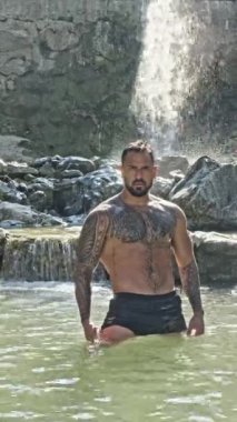 Dağ nehri şelalesinin altındaki adam, gül kollarını kaldırdı ve su sıçratmaktan zevk alıyor. Doğada sıcak bir yaz. Su doğasında rahatla. Kaslı gövdeli seksi adam, su sıçratmaktan hoşlanıyor. Doğada yıkanmak.