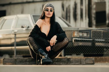 Güneş gözlüğü ve bandana takan seksi hippi kadın, siyah çizme ve ceket. Küstah moda kızı. Hip-hop kızı. Moda kadını dışarıda. Sokakta oturan şık, genç, seksi hippi kadın modeli.