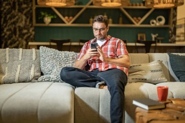 Evde oturan cep telefonu olan yakışıklı bir adam. Amerikalı bir adam oturma odasındaki koltukta otururken telefon kullanıyor. Mobil cihazlarda çalışan insanlar kavramı. Evde akıllı telefonla rahatlamak.