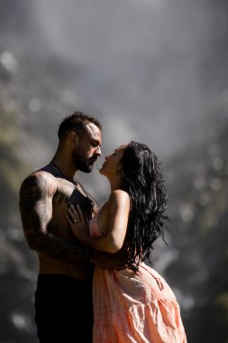 Şehvetli bir çift şelalenin yanında öpüşüyor. Tutkulu çift yaz doğasına karşı sarılıyor. Kırsalda romantik aşıklar. Seksi çiftin moda portresi muhteşem manzarada birbirine sarılıyor.