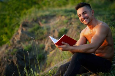 Çıplak vücutlu, açık havada kitap okuyan seksi, mutlu bir adam. Yakışıklı bir entelektüel. Seksi vücut okuma kitabı olan inek ya da zeki bir erkek model. Parktaki çimlerin üzerinde kitap tutan adam.