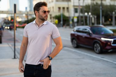 Güneş gözlüğü ve gömlek takan şık bir adam. Dışarıdaki yakışıklı adam portresi. Açık havada şık bir erkek modelin portresi. Moda erkeği sokakta gökdelenin yakınında poz veriyor. Şehir tarzı modaya uygun bir adam.