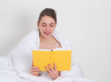 Genç kadın yatakta kitap okuyor. Yatakta kitap okuyan güzel bir kız. Rahatlatıcı bir kavram. Kız yatak odasında kitap okuyor. Dinlenme, rahatlık ve rahatlık. Yatakta kitap okuyan mutlu genç bir kadın.