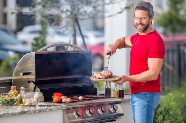 Barbekü. Barbekü için tişörtlü orta yaşlı bir adam. Kızartma ve ızgara yemek. Adam mutfak aletlerini barbeküde tutuyor. Açık havada et kızartıyoruz. Barbekü ve ızgara. Arka bahçede et pişiriyorum.