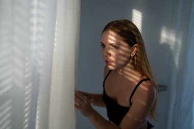 Pencere perdelerinden bakan romantik kadın. Pencere perdelerinden gölgelerde bir kadının portresi. Kız oturma odasındaki pencereye dalmış bakıyor. Perdelerin gölgesi olan kadın portresi.