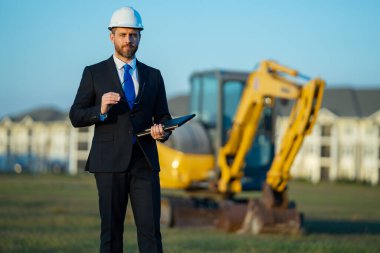 Başarılı bir inşaat işletmesi sahibi. Kazıcının yanındaki kask ve takım elbiseli inşaat işçisi. İnşaat alanının önünde kendine güvenen bir inşaat sahibi. İnşaat mühendisi.