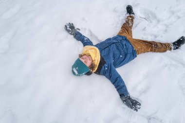 Kış doğasında karda yatan bir çocuğun portresi. Karlı bir kış parkında oynayan örgü şapkalı bir çocuk. Çocuk kışın karla oynuyor. Çocuk karlı ormanda oynuyor. Çocukla kış tatili