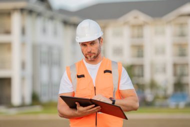 Dosyası olan mühendis, bina teftişi. Miğferli inşaat işçisi yeni bir ev inşa etti. Mühendis, inşaat üniforması ve başlık giyiyor. Foreman açık hava portresi