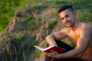 Açık havada kitap okuyan çıplak vücutlu seksi bir adam. Yakışıklı bir entelektüel. Seksi vücut okuma kitabı olan inek ya da zeki bir erkek model. Parktaki çimlerin üzerinde kitap tutan adam.