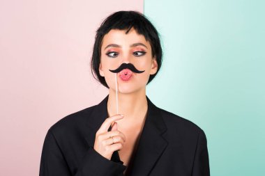 Siyah peruklu genç bir kadın sahte bıyığı pozitif ve mutlu yapıyor. Komik kız gülüşü. Genç transseksüel kadın. Komik kadın suratı. Çekici, suratını buruşturan komik kızın portresi. Duygu kızı.