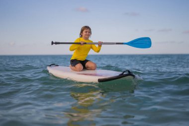 Çocuk SUP yatılı. Çocuk kürek çekiyor ya da kürek çekiyor. Sağlıklı yaz yaşam tarzı. Yaz Suyu sporu. Yaz tatili macerasında SUP sörf turu. Kid Boy raket suyu ile yüzüyor.