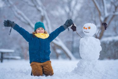Komik Çocuk kışın kardan adam için kartopu yapıyor. Çocuk karda oynuyor. Açık havada aktif kış oyunları. Çocuk karla oynarken eğleniyor. Çocuklar için açık havada kış tatili. Güneşli kış gününde çocuk oyunu