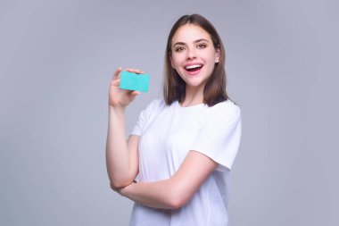 Yüzünün yanında kredi kartı tutan beyaz tişörtlü genç bir kadın. Gri arka planda izole edilmiş. Zeka ya da banka kartını öde. Banka kartı. Plastik kredi kartı