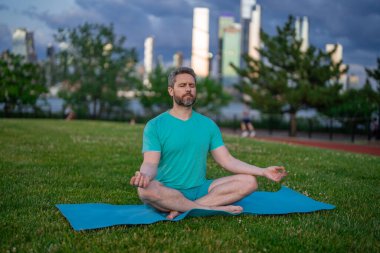 Nilüfer çiçeği içinde meditasyon yapan yaşlı bir adam açık havada çimlerde poz verir. Sporcu şehir parkında yoga yaparken asana meditasyonu yapıyor. Atletik erkek meditasyonu, rahatlama, sağlıklı yaşam tarzı kavramı. Açık havada meditasyon.