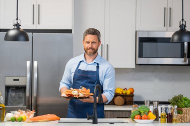 Ev yapımı modern mutfakta balık somonu hazırlayan önlüklü adam. Yakışıklı adam mutfakta taze balık somon balığı, yengeç, karides ve ıstakoz pişiriyor.