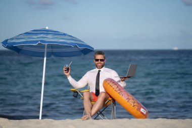 Yaz tatillerinde başarılı bir iş adamı. Komik iş adamı deniz sahilinde yaz kokteyli içip dizüstü bilgisayar kullanıyor. Yaz işi konsepti. Sıcak yaz. Çılgınca. Uzak çevrimiçi çalışma