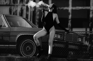 Beautiful sexy women vaping outdoor. Fashion model sitting near retro car and smokes electronic vape outdoor. Stylish girl smoking an e-cigarette. Modern girl smoking vape. Vaping concept clipart