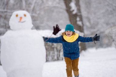 Çocuk kışın kardanadamla oynuyor. Kış günü havuç burunlu bir kardan adam. Kardan adam yapan çocuk. Kar parkında kardan adam yapan küçük çocuk. Karlı kış parkında kardan adamı kucaklayan çocuk