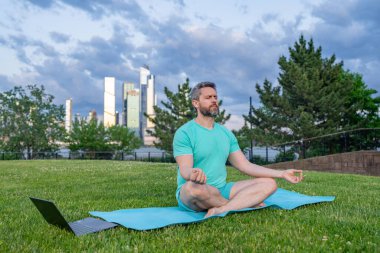 Nilüfer çiçeği içinde meditasyon yapan bir adam dışarıda çimlerde poz verir. Spor adamı, şehir parkında yoga yapıyor. Atletik erkek meditasyonu, rahatlama, aktif sağlıklı yaşam tarzı kavramı. Açık havada meditasyon.