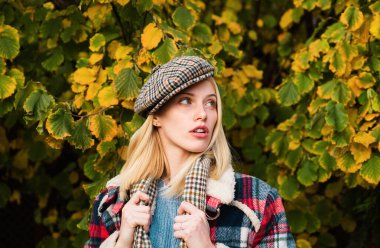 Beautiful femme wearing plaid scarf and cap in parisian style. Autumnal fashion concept. Beautiful romantic blonde girl standing at fall yellow autumnal leaves and looks dreamy clipart