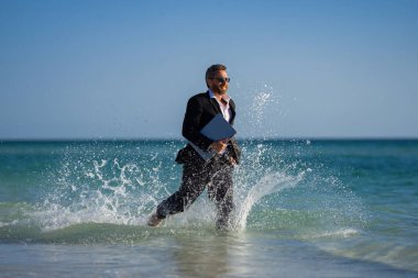 Serbest iş, online iş. Yaz işi. Takım elbiseli iş adamı dizüstü bilgisayarı tutuyor ve denizde koşuyor. Suda dizüstü bilgisayarı olan takım elbiseli komik bir iş adamı. Uzaktan kumanda. Çılgın serbest çalışan.