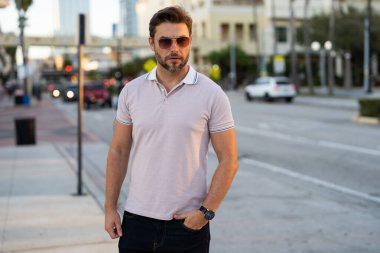 Şehir merkezinde sıradan, yakışıklı bir manken. İyi giyimli, gözlüklü ve polo pozu veren bir adam. Erkek model. Şehirde şık bir adamın portresi. Açık hava moda portresi