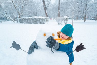 Soğuk bir kış gününde, kış parkında heyecanlı bir çocuk. Kışın karla oynayan sevimli bir çocuk. Kardan adamla komik çocuk portresi. Noel 'de kış aktiviteleri. Parkta kardan adam yapan çocuk