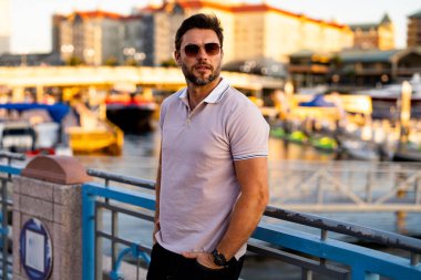 Güneş gözlüğü ve gömlek takan şık bir adam. Dışarıdaki yakışıklı adam portresi. Açık havada şık bir erkek modelin portresi. Moda erkeği sokakta gökdelenin yakınında poz veriyor. Şehir tarzı modaya uygun bir adam.