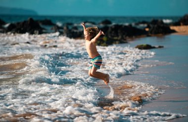 Okyanus sahilinde oynayan bir çocuk. Deniz tatilinde dalgalara atlayan bir çocuk. Yaz tatili boyunca egzotik adanın tropik sahillerinde koşan küçük çocuk.
