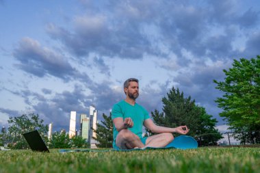 Nilüfer çiçeği içinde meditasyon yapan yaşlı bir adam açık havada çimlerde poz verir. Sporcu şehir parkında yoga yaparken asana meditasyonu yapıyor. Atletik erkek meditasyonu, rahatlama, sağlıklı yaşam tarzı kavramı. Açık havada meditasyon.
