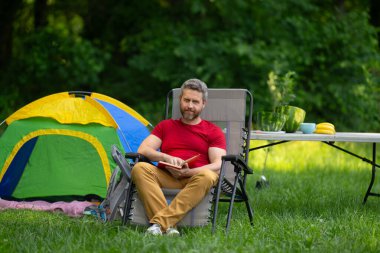 Adam doğada kamp yapmayı seviyor. Doğaya yaz gezisi. Orman kampında dinlenen bir turist. Kamp gezisi. Yürüyüşten sonra çadırın yanında dinleniyor. Barışçıl bir kamp alanında özgürlük tatili. Yazın piknikteki adam