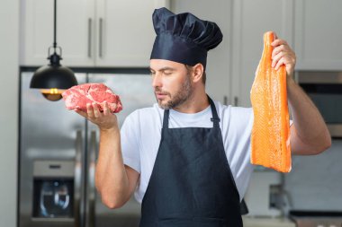 Aşçı portresi. Mutfakta balık, et, somon ve sığır eti var. Önlük, şef üniforması ve aşçı şapkası takan bir adam. Reklam için çiğ et ve somon filetosu.