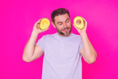 Adam pembe arka planda izole edilmiş bir avokado tutuyor, stüdyo portresi. Avokado. Sağlıklı görünüş. Yakışıklı bir adamın portresi Avokado 'yu yüzüne yakın tutar.