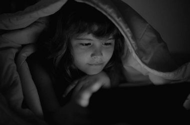 Çocuk oyuncağı bağımlılığı ve uykusuzluk. Sosyal medyası olan çocuklar. Yatakta uzanmış çocuk, karanlık odada tablet çalıyor, Blacket 'in altında ışık var. Çocuğun tabletten çizgi film izlemesini kapat.