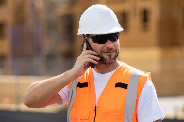 Bina konsepti. İnşaatçı ustabaşı. İnşaat alanında miğferli beyaz adam inşaat işçisi. Çalışanlar telefon kullanıyor, inşaatçılar işe ara veriyor. Cep telefonlu mimar