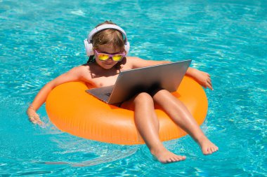 Yaz seyahati ve iş konsepti. Çocuk yaz havuzunda dizüstü bilgisayarla çalışıyor. Bilgisayar kullanan, havuz başında çalışan küçük bir serbest çalışan.