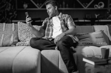 Serbest çalışan bir iş adamı cep telefonu kullanıyor sosyal medyayı kontrol ediyor ya da evde mesajlaşma sohbeti yapıyor. İnternette evden çalışıyor. Adam oturma odasında sıkı çalışma gününden sonra dinleniyor. Evdeki koltukta cep telefonu kullanan bir adam.