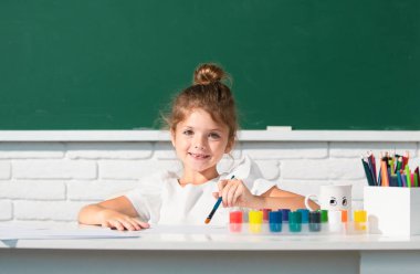 Resim dersi, çizim sanatı. Kız çocuğu sınıfta resim çiziyor, masada oturuyor, okul karatahtasında eğleniyor.