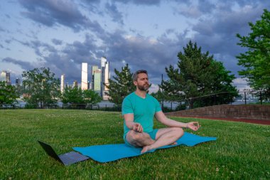Nilüfer çiçeği içinde meditasyon yapan yaşlı bir adam açık havada çimlerde poz verir. Sporcu şehir parkında yoga yaparken asana meditasyonu yapıyor. Atletik erkek meditasyonu, rahatlama, sağlıklı yaşam tarzı kavramı. Açık havada meditasyon.