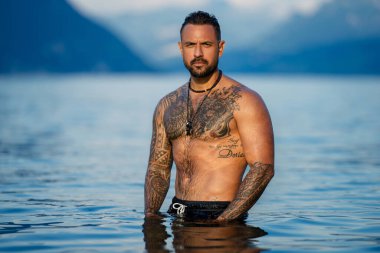 Seksi yaz erkeği. Suda ıslak kaslı erkek vücudu. Çıplak seksi erkek vücudu. İnsan yaz doğasında suya su sıçratır. Yaz plajında poz veren formda bir adam. Dağ gölünde su sıçratan güçlü bir adam. Yaz gezisi