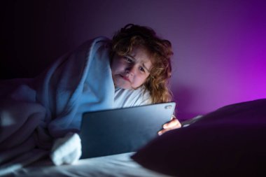 Çocuk yatakta uzanıyor ve karanlık odada tablet üzerinde internette sörf yapıyor. Geceleri Tablet okuyan bir çocuk. Yatmadan önce pijamalı bir çocuk tablet kullanıyor. Çocuk yatakta tabletini izliyor.