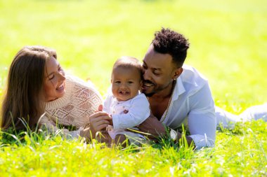 Çok ırklı ebeveynler ve parkta melez bir bebek. Çok ırklı aile portresi. Bahçede melez bir bebek var. Çeşitli ailelerden. Afrika kökenli Amerikalı baba beyaz anne ve bebeği çimenlerde yatıyor.