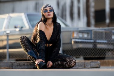 Garaj duvarında poz veren seksi, şehvetli bir kadın. Hippi tarzı giyinmiş hippi bir genç kadın. Siyah giysili şık bir kız. Güzel seksi kadın eski arabanın yanında oturuyor.