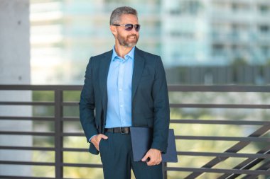 Başarılı bir iş adamı dışarıda dizüstü bilgisayarla dolaşır. Kendine güvenen iş adamı dizüstü bilgisayarı dışarıda tutuyor. Takım elbiseli, olgun, serbest çalışan bir iş adamı. Başarı işi. Takım elbiseli zengin iş adamı