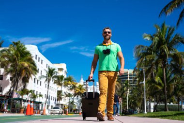 Bir gezgin Miami 'nin güneşli sokaklarında yürüyor. Bir erkek gezgin, tropik bir şehirde yaz tatilinin tadını çıkarır. Bavullu bir gezgin turist şehir manzarası. İnsan gezgin şehirde yürür