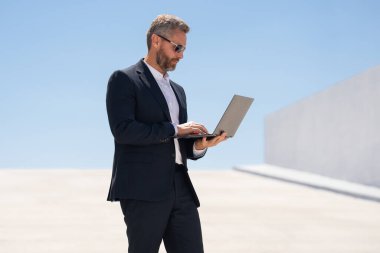 İş adamı dışarıda dizüstü bilgisayar kullanıyor. Dizüstü bilgisayarda çalışan başarılı bir adam. Dizüstü bilgisayarı olan kendinden emin bir iş adamı. İnternette iş. Şehir işadamı dışarıda dizüstü bilgisayardan verileri kontrol ediyor. Takım elbiseli adam internette çalışıyor.