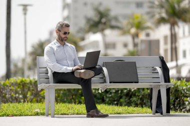Kendine güvenen bir iş adamı bankta dizüstü bilgisayarı ve parktaki telefonuyla oturuyor. Zengin iş adamı. Olgun iş adamı tatilin tadını çıkarıyor. İş adamı parktaki bankta mola veriyor. Kentsel iş